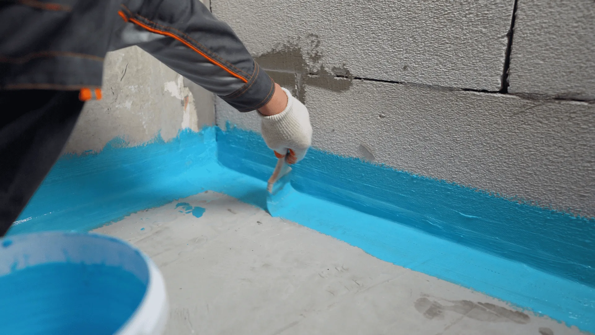 Worker brushing blue waterproofing membrane along the base of a wall