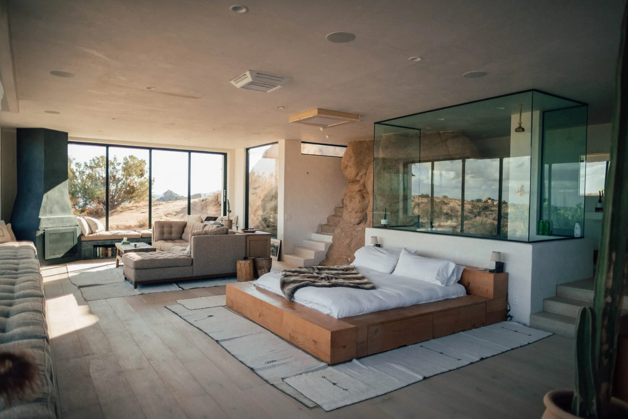 Open-plan modern bedroom and lounge with a timber platform bed, a glass-walled bathroom and desert views
