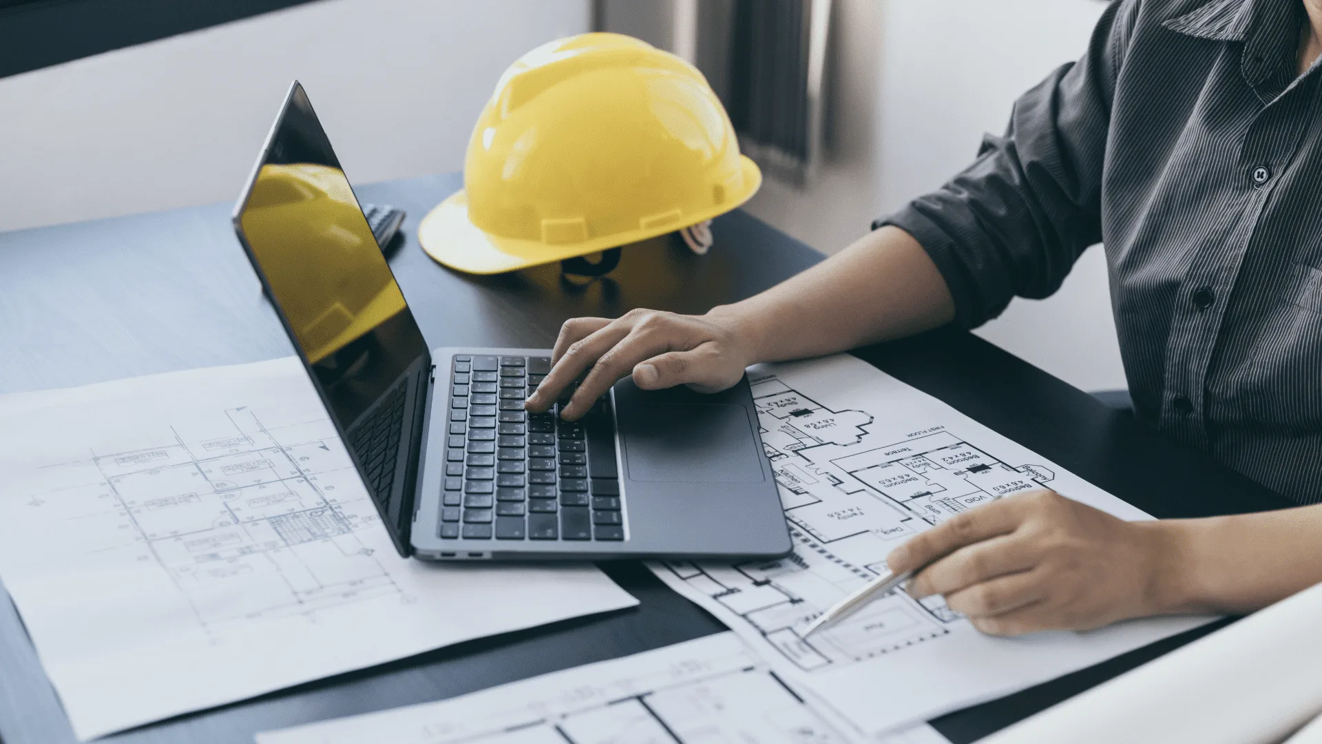 Estimator working through blueprints on a laptop beside a hard hat