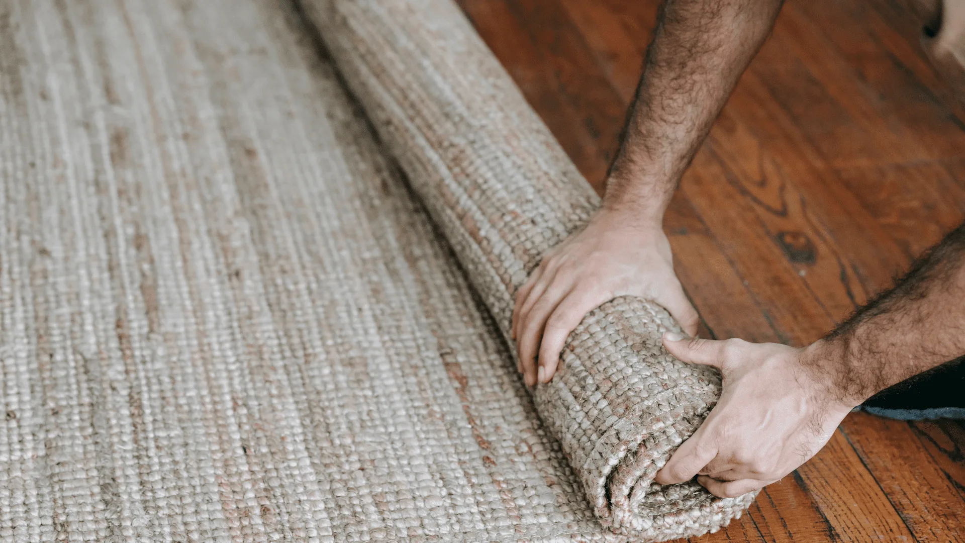 Hands unrolling a woven carpet across a wood floor
