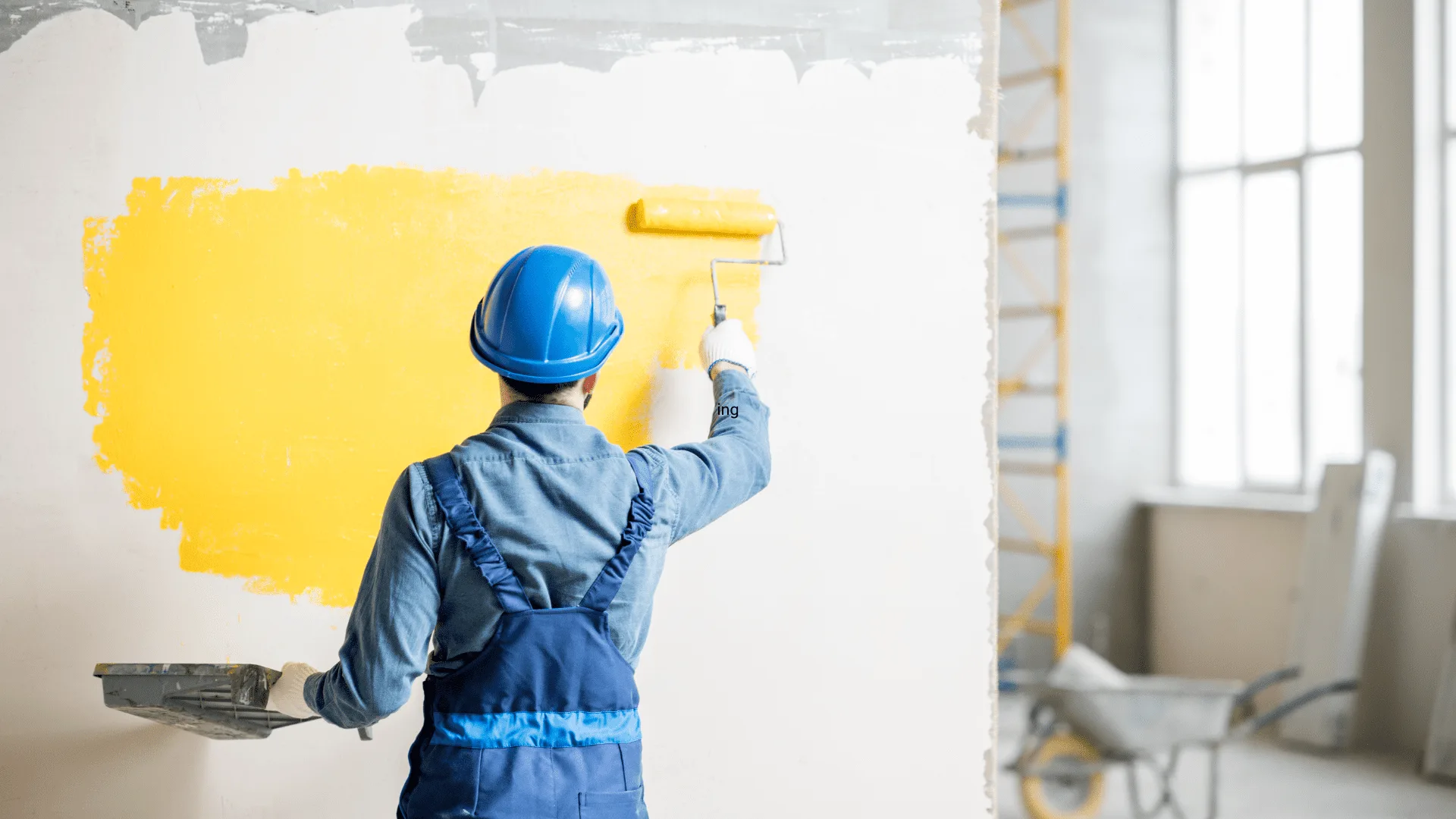 Painter in a hard hat rolling yellow paint onto an interior wall