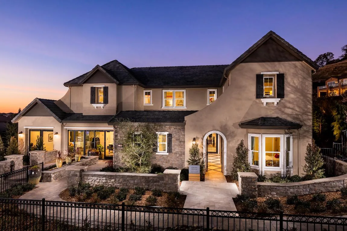 Two-storey stucco and stone house front elevation at dusk with warm lit windows and an arched entry