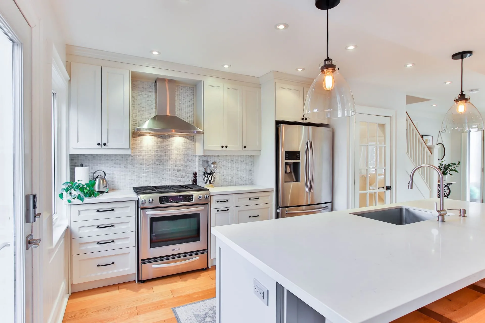 White shaker kitchen with a mosaic backsplash, a stainless range hood and a quartz island under glass pendant lights